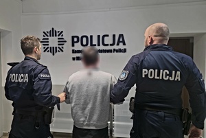 Dwaj policjanci przytrzymujący zatrzymanego mężczyznę na tle wewnętrznej ściany z logiem Policji.