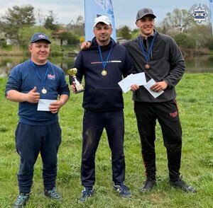 Trzech mężczyzn z medalami na szyjach nad brzegiem zbiornika wodnego, jeden z nich w rękach trzyma mały puchar
