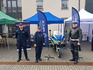 Na tle budynku namiot wystawienniczy w kolorze granatowym, trójka policjantów oraz motocykl policyjny