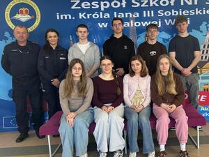 Dwójka policjantów pozujących do zdjęcia wraz z młodzieżą na tle ściany z nazwą szkoły