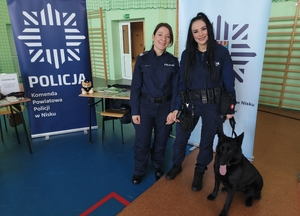 Wewnątrz sali dwie policjantki na tle stoiska wystawienniczego KPP w Nisku, obok pies służbowy