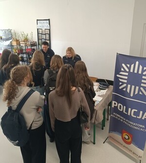 Zdjęcie z góry, w sali stoisko ekspozycyjne przy którym siedzą dwie policjantki oraz stoi oczekująca grupa młodzieży