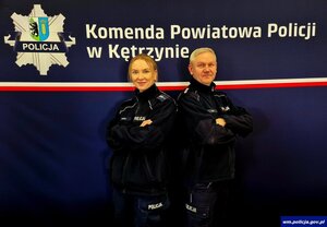 Dwójka uśmiechniętych policjantów na tle banneru Komendy Powiatowej Policji w Kętrzynie