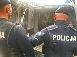 Widziani z tyłu dwaj policjanci przy otwartych drzwiach z pomieszczeniem dla zatrzymanych radiowozu typu bus