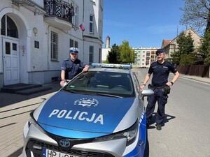 Dwaj policjanci po obu stronach radiowozu na tle kamienicy