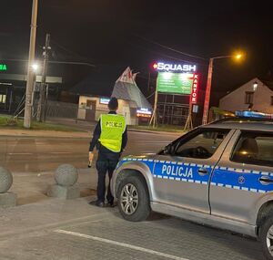 W scenerii  nocnej widziany z tyłu policjant stojący na krawędzi chodnika obok radiowozu