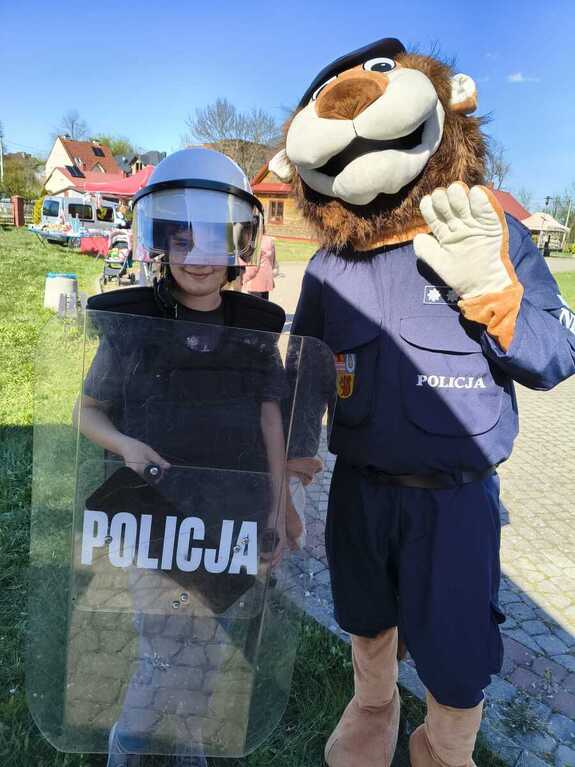 W otwartej przestrzeni dziewczynka ubrana w kasj policyjny z tarczą ochronną pozująca z maskotka policyjną w postaci lwa