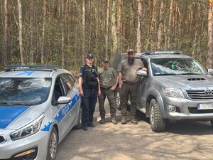 Na leśnej polanie obok radiowozu i samochodu terenowego policjant i dwaj strażnicy leśni