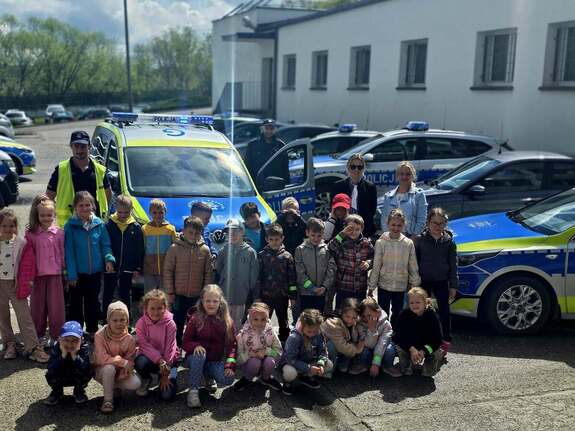 Na zewnątrz budynku grupa przedszkolaków pozująca z policjantem do zdjęcia wokół radiowozu