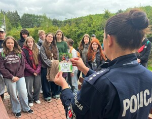 Widziana z tyłu policjantka prowadząca wykład dla zgromadzonej na tle zieleni młodzieży