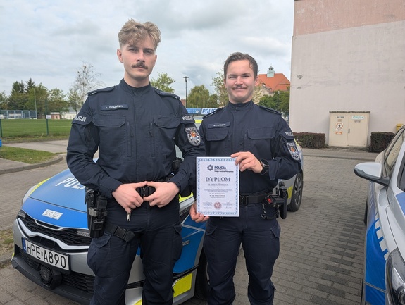 Dwóch policjantów obok radiowozów na tle budynku, z których jeden w rękach trzyma dyplom