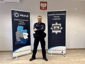 Wewnątrz pomieszczenia uśmiechnięty policjant obok bannerów Komendy Policji w Sieradzu