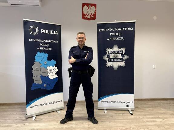 Wewnątrz pomieszczenia uśmiechnięty policjant obok bannerów Komendy Policji w Sieradzu