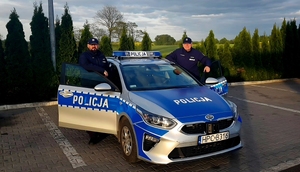 Dwaj policjanci po obu stronach radiowozu w otwartych drzwiach, w tle zieleń