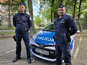 Dwaj policjanci przed radiowozem na tle zieleni