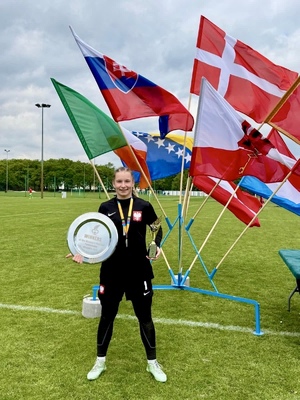 uśmiechnięta kobieta z trofeami sportowymi w rękach na tle różnych flag na boisku sportowym