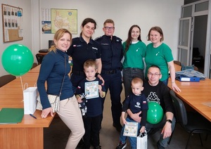 W pomieszczeniu dwie policjantki, trzy kobiety w zielonych tiszertach, mężczyzna oraz dwóch małych chłopców trzymających w rękach dyplomy