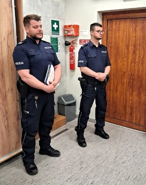 Zbliżenie dwóch policjantów w rogu pomieszczenia, w tle gaśnica oraz tablice ostrzegawczo-informacyjne