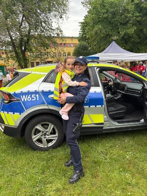 Uśmiechnięta policjantka trzymająca na rękach dziecko na tle radiowozu i zieleni miejskiej