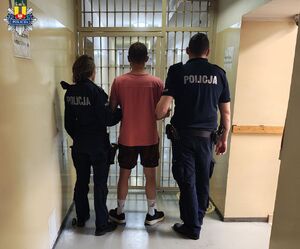 Przed kratą aresztu widziani z tyłu dwójka policjantów przytrzymująca mężczyznę w krótkich spodenkach