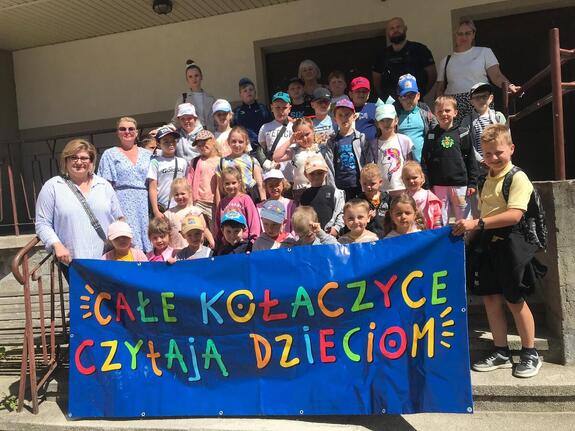 Na schodach wejściowych grupa dzieci wraz z wychowawczyniami i policjantem trzymająca transparent "Całe Kołaczyce czytają dzieciom"