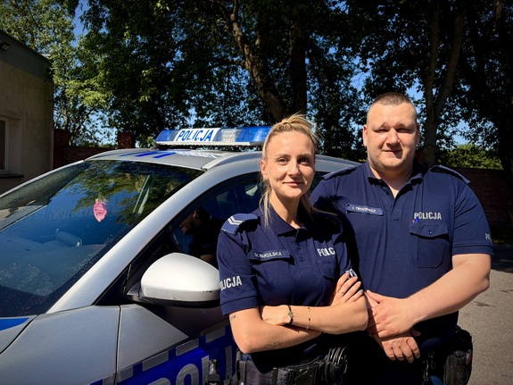 Uśmiechnięci policjantka i policjant na tle radiowozu i zieleni drzew
