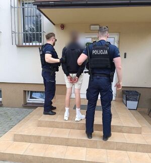 Widziani z tyłu dwaj policjanci przed wejściem do siedziby Policji przytrzymujacy mężczyznę ze skutymi rękami