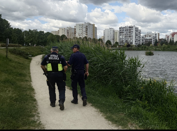 Widziani z tyłu policjant i strażnik miejscy idący alejką wzdłuż akwenu, w tle bloki  mieszkalne