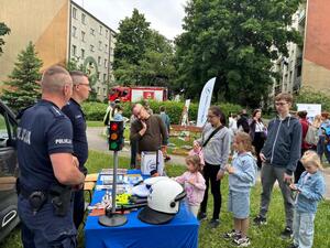 Na tle zieleni i osiedla mieszkaniowego stolik wystawienniczy przy nim dawaj policjanci oraz dzieci z opiekunami