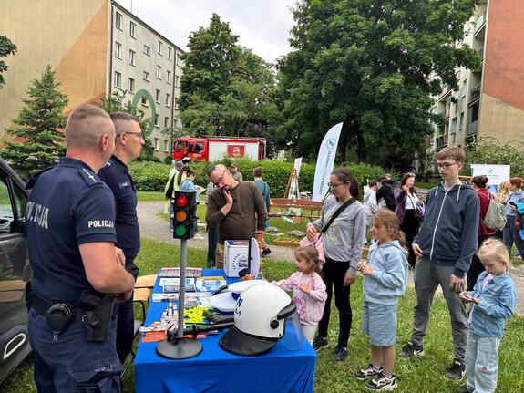 Na tle zieleni i osiedla mieszkaniowego stolik wystawienniczy przy nim dawaj policjanci oraz dzieci z opiekunami