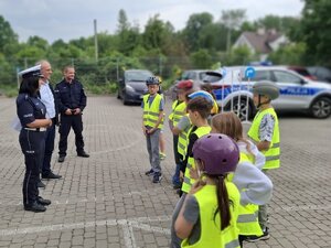 Na placu grupka dzieci w kaskach ochronnych, mężczyzna oraz dwójka policjantów