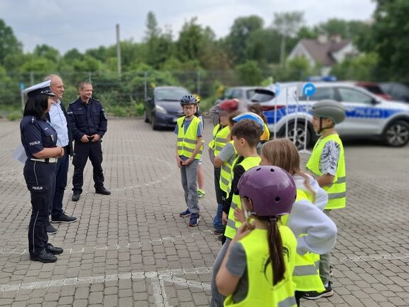 Na placu grupka dzieci w kaskach ochronnych, mężczyzna oraz dwójka policjantów