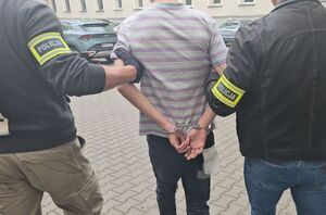 Zbliżenie pleców mężczyzny w koszulce na dziedzińcu komendy przytrzymywanego przez dwóch policjantów