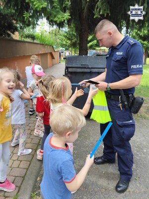 Policjant rozdający dzieciom opaski odblaskowe na tle zieleni dziedzińca