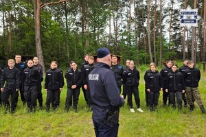 Na tle lasu policjant oraz grupa młodzieży ustawiona w dwuszeregu