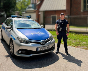 Policjant obok radiowozu na tle zabytkowych drewnianych zabudowań