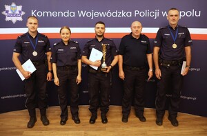 Piątka policjantów z dyplomami i pucharami w rękach na tle ścianki Komendy Wojewódzkiej Policji w Opolu