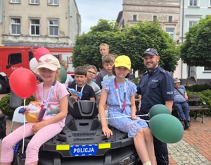 Uśmiechnięty policjant z grupą dzieci siedzących na quadzie na tle wozu strażackiego