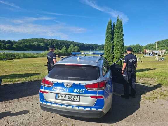 widziani z tyłu dwaj policjanci po obu stronach radiowozu na terenach zielonych w tle zbiornik wodny