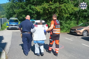 Na skraju skweru miejskiego policjant i ratownik medyczny prowadzący pod ręce starszą kobietę