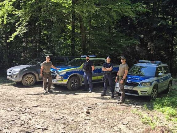 Na skraju leśnej polany dwa radiowozy, samochód terenowy straży leśnej oraz dwóch policjantów i dwóch strażników leśnych