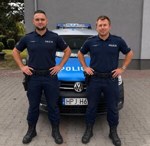 Dwaj uśmiechnięci policjanci pozujący do zdjęcia na tle radiowozu