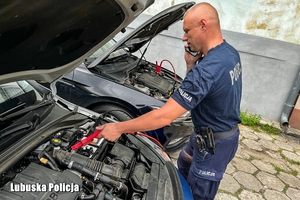 Policjant z telefonem w ręku przy dwóch samochodach z podniesionymi pokrywami silnika uruchamiający je na tzw. kable