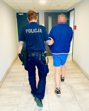 Widziani od tyłu: policjant prowadzący korytarzem mężczyznę w granatowej kurtce