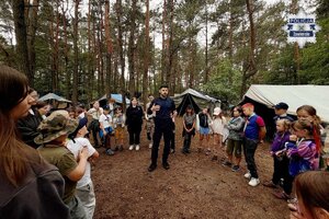 Policjant na leśnej polanie prowadzący pogadankę dla zebranych dzieci i młodzieży, w tle namioty harcerskie