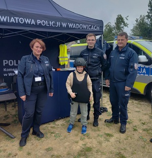 Pozująca do zdjęcia przed namiotem wystawienniczym trójka policjantów oraz chłopiec w hełmie i kamizelce balistycznej