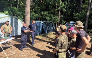 Dwaj policjanci prowadzący pogadankę dla harcerzy zgormadzonych na leśnej polanie, w tle namioty