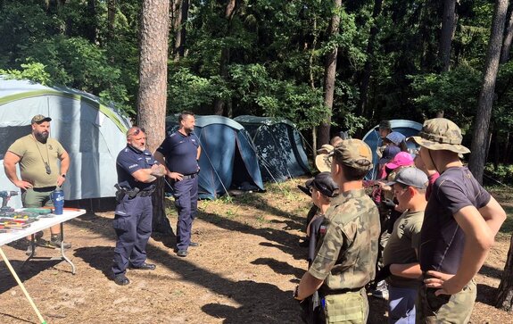 Dwaj policjanci prowadzący pogadankę dla harcerzy zgormadzonych na leśnej polanie, w tle namioty