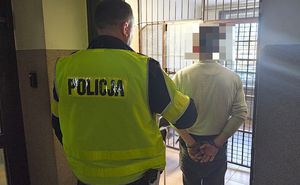 Widziani z tyłu w korytarzu przed kratami aresztu: policjant i mężczyzna, ze skutymi z tyłu kajdankami rękami