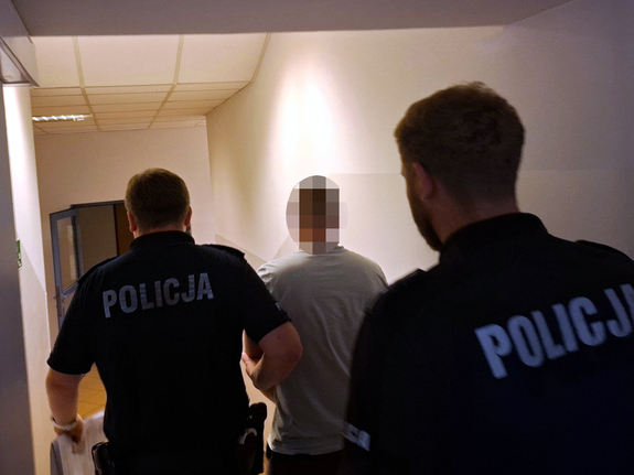 Widziani z tyłu dwaj policjanci prowadzący korytarzem zatrzymanego